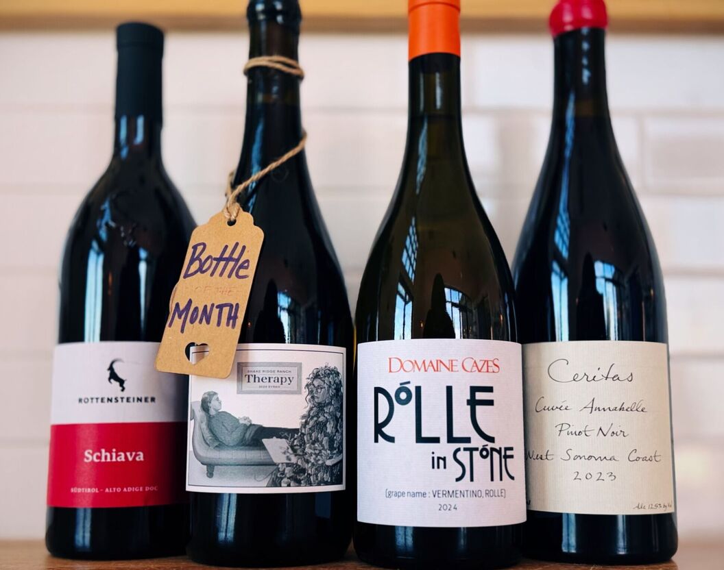 Four featured wine bottles: Rottensteiner Schiava, BXT Therapy Syrah (Bottle of the Month), Domaine Cazes Rôlle Vermentino, and Ceritas Cuvée Annabelle Pinot Noir, displayed on wooden bar top.