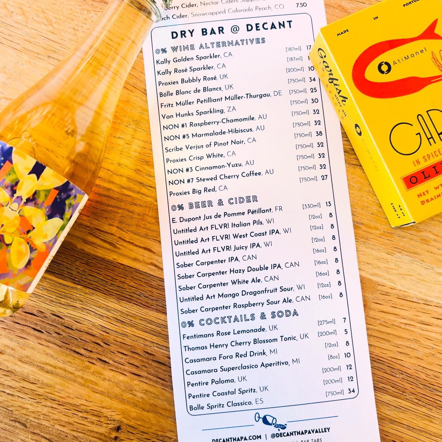 DECANT's Dry Bar menu featuring 0% wine alternatives, beers, ciders, cocktails and sodas, displayed on wooden table with a Kally wine bottle and yellow flowers